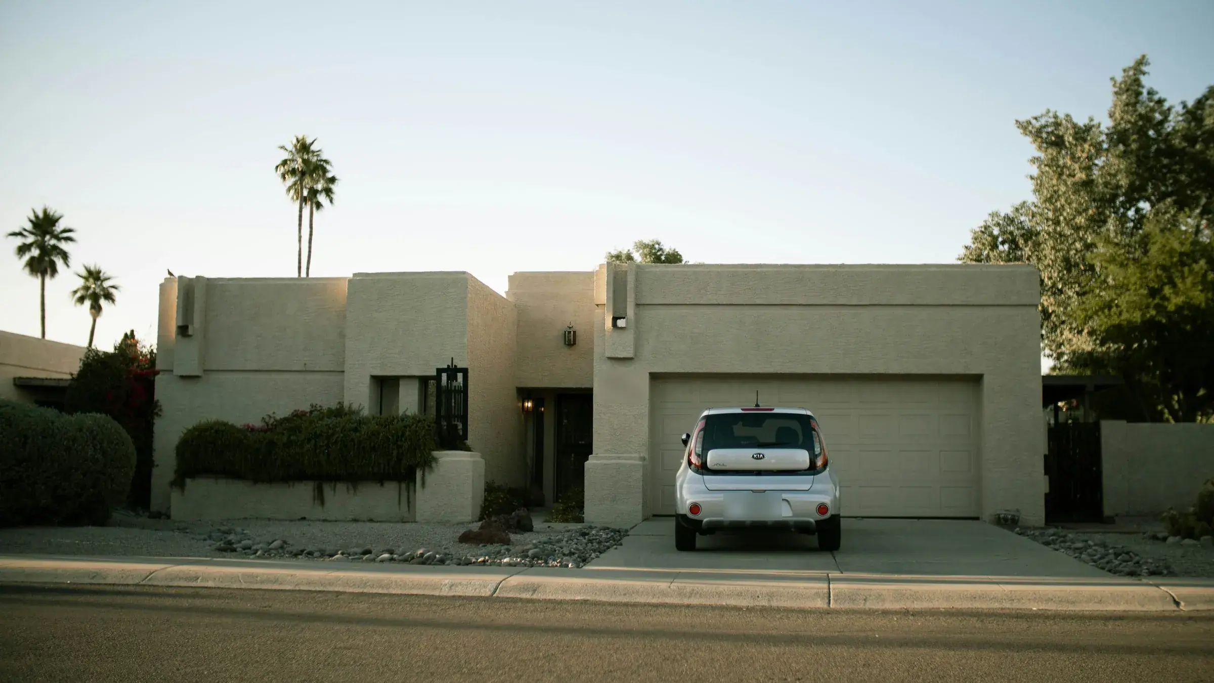 A flat-roofed stucco house in low desert light, a silver car parked on the driveway in front of its two-car garage door, gravel and boulders across the front yard.